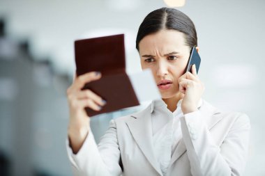 Nervous businesswoman looking at her passport and flight ticket while talking by smartphone in airport clipart