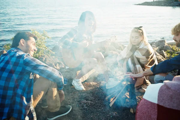 Several happy friends sitting around campfire on summer evening by seaside