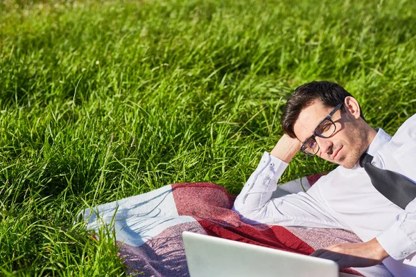 严肃的年轻商人躺在草坪上的笔记本电脑和分析在线数据 — 图库照片