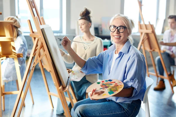 混合油絵具やイーゼルの前に座っている絵筆のパレットを持つ成熟した女性の笑顔 — ストック写真