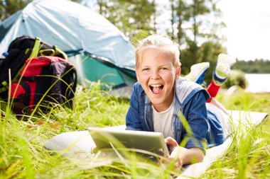 Neşeli çocuk çimenlerin üzerinde rahatlatıcı ve net tarama yaparken kameraya bakarak tablet ile