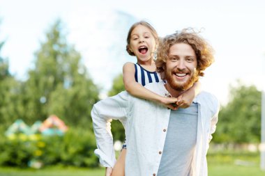 Sırtında küçük kız ve her iki seyir alarak yürüyüş sırasında kamera tutan dişlek gülümseme ile neşeli genç baba