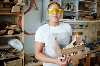 Happy female worker with wooden toolbox wearing protective eyeglasses during work clipart