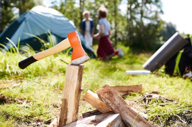Ax in piece of firewood prepared for campfire with scouts by tent on background clipart