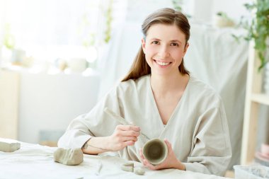 Kendi kendine yapılan kil kupa ve süs eşyaları kesme ve kamera onun işyeri tarafından bakarak handtool tutan gülümseyen kız