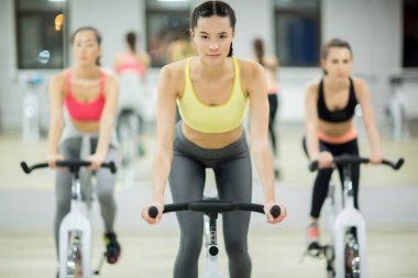 Genç sportif kadın fitness Center'da eğitim bisiklet önünde eğitmen ile sürme