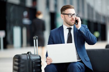 Ciddi genç Ajan veya komisyoncu ile salonda için uçak beklerken smartphone tarafından istemciye konuşan laptop