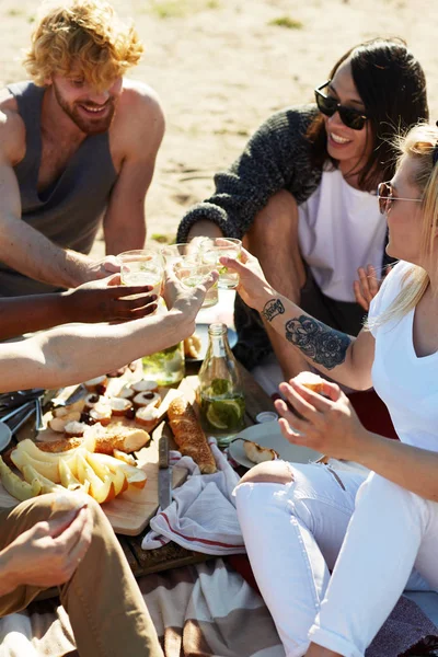 Several young friends toasting with glasse sof lemonade over snack during picnic on beach