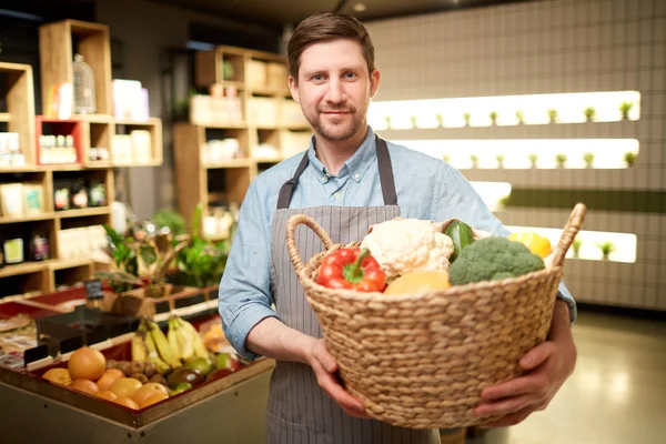 Happy supermarket assistant holding big basket with fresh vegetables during work