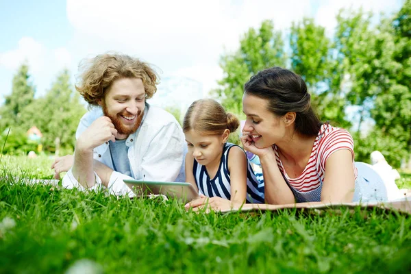 当代家庭的父亲 母亲和女儿的平板上躺在绿色的草坪上 放松和观看视频在网上 — 图库照片