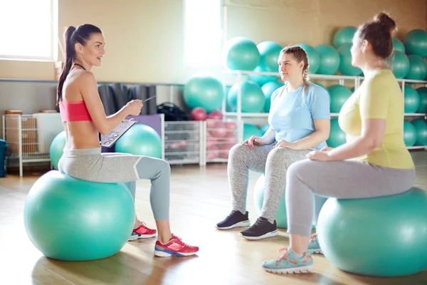 Young fitness instructor and her group sitting on green fitballs in gym and discussing which exercises they are going to do