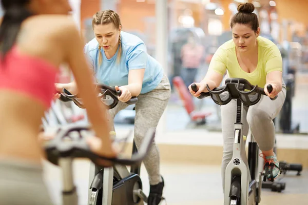 胖女孩在运动装坐在健身自行车和自行车在健身房锻炼与他们的教练 — 图库照片