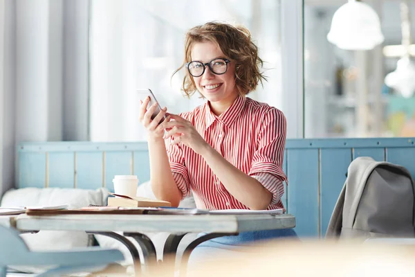 カフェのテーブルに座って ソーシャル ネットワークの友人と彼女の写真を共有しながらカメラを見て幸せな女の子 — ストック写真