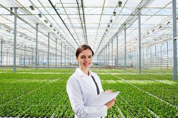 Young successful biologist in whitecoat on background of large green plantation in glasshouse