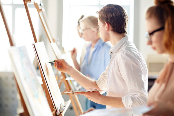 Creative guy looking at picture on easel of teacher while painting at lesson in studio of arts