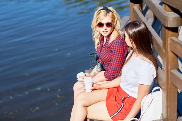 两个友好的女孩用塑料杯咖啡坐在木凳上的水 而放松在阳光明媚的一天 在公园 — 图库照片