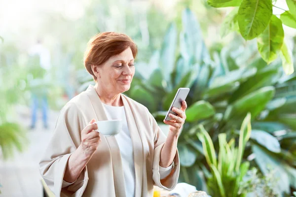 オランジェリーの緑の植物の中朝のお茶をしながらスマート フォンで幸せな成熟した女のテキスト メッセージ — ストック写真