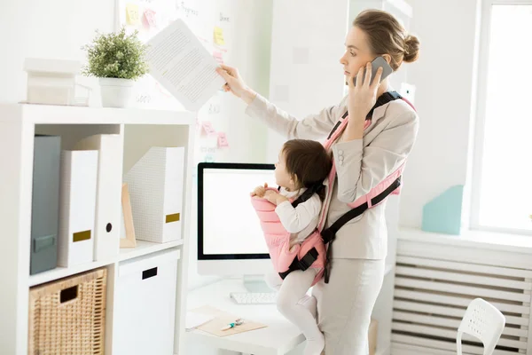 Young business mother with documents talking by smartphone in office with her baby in carrier