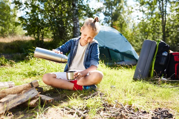 若々しい少年の夏の日を楽しんで彼のテントの背景の上で焚き火しながらマグカップでお茶を注ぐ — ストック写真