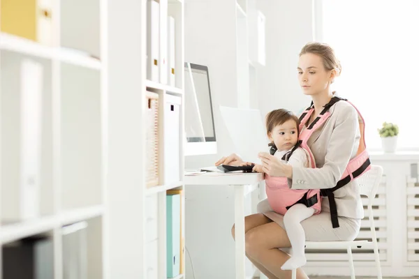Adorable baby in pink carrier sitting close to her busy mother looking though financial documents in front of computer in office