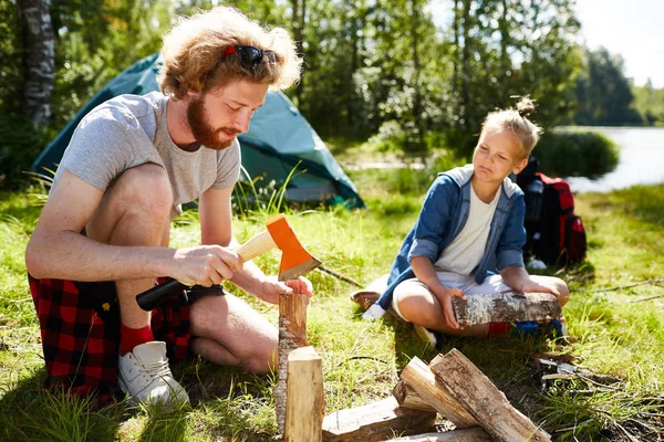 彼の息子の旅行の間に夏の日の焚き火用の薪をカットする方法を示す斧を持った若い男 — ストック写真