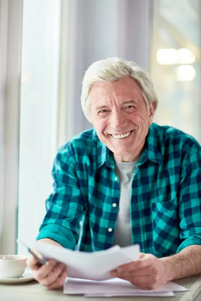 Cheerful aged man with papers looking at camera while spending time in cafe