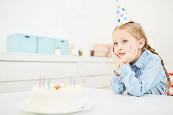 幼稚園でロウソクでケーキの前のテーブルに座って誕生日キャップで物思いにふける女の子 — ストック写真