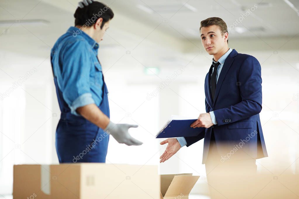 Young businessman with document pointing at open box while talking to relocation service staff in new office