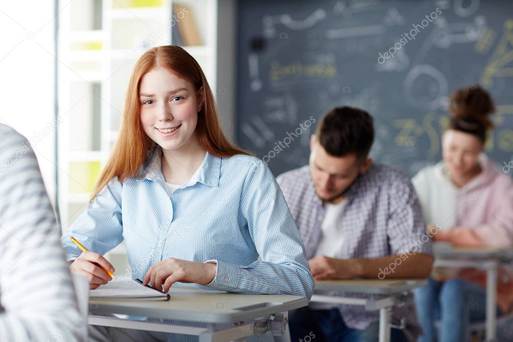 Smiling girl looking at camera while writing final exam test at lesson between her classmates