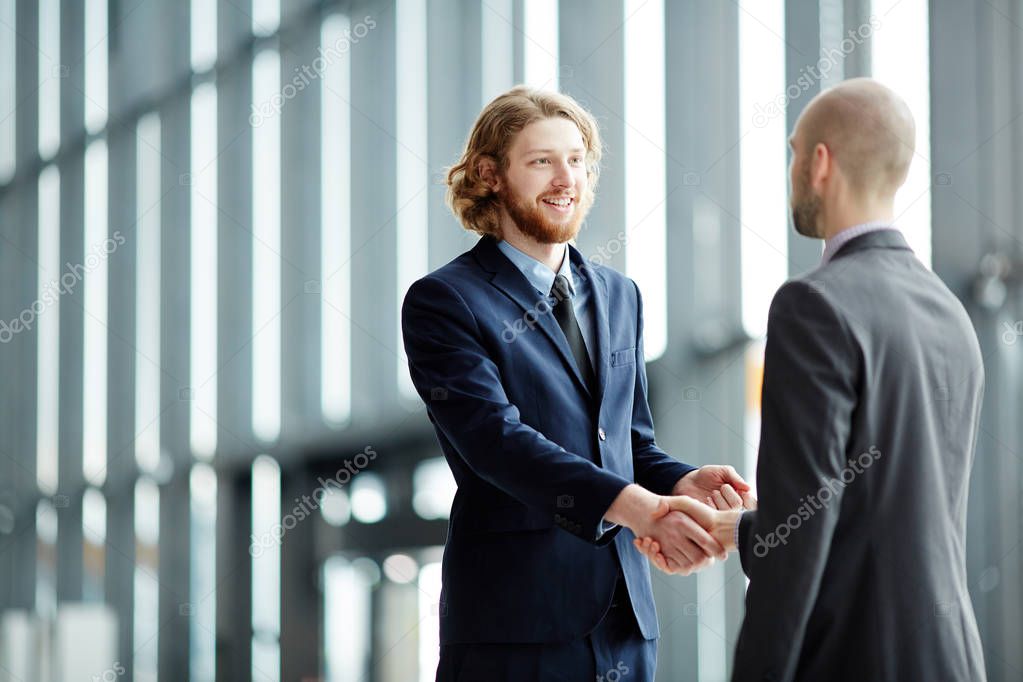 Two young businessmen in suits greeting one another by handshake after confirming deal