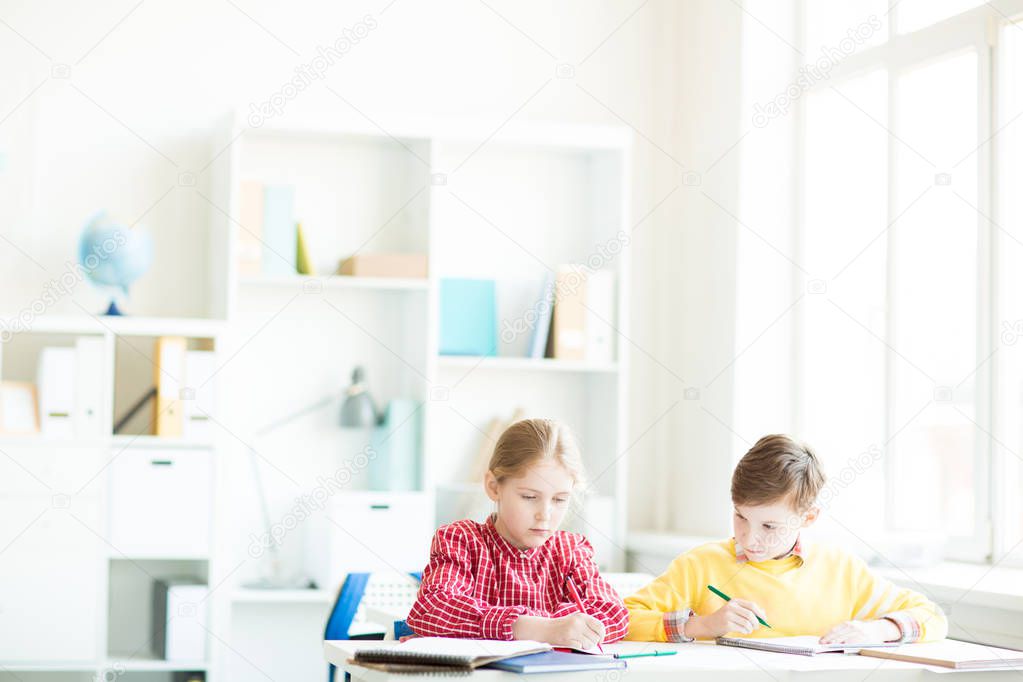 Two little classmates sitting by desk and drawing or making notes in their copybooks at lesson