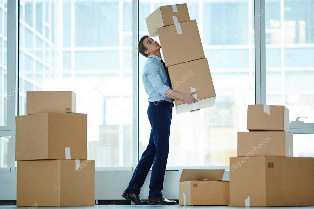 Young businessman carrying huge stack of big boxes with office supplies during relocation to modern business center