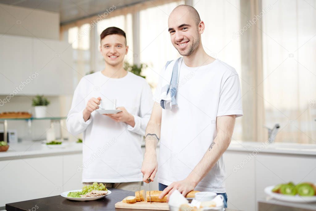 Happy young man cutting baguette for breakfast while standing by table in the kitchen