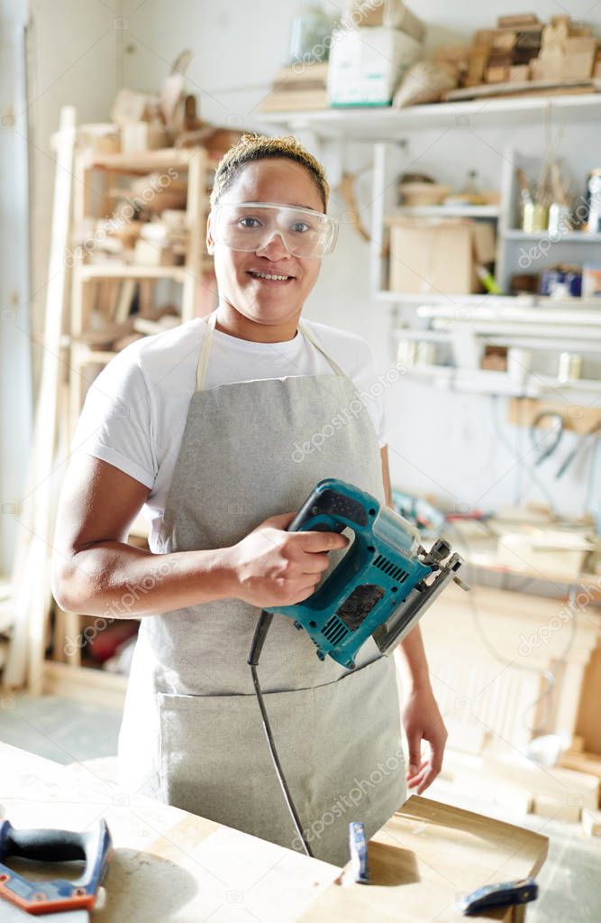 Female carpenter with electric fretsaw looking at camera through protective eyeglasses in workroom
