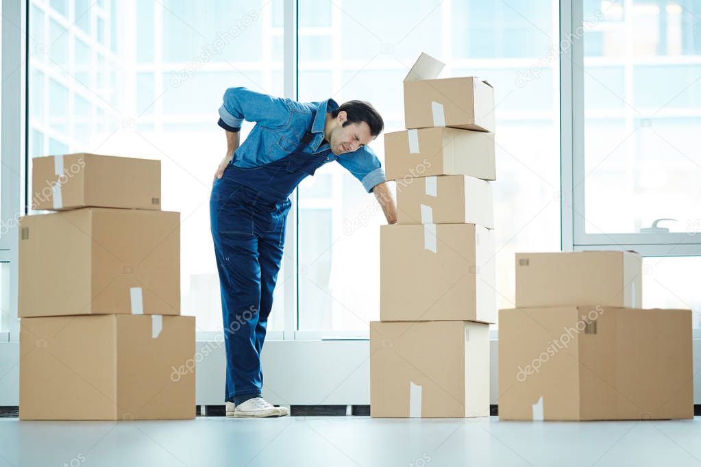 Young relocation service staff touching his back and leaning by stack of packed boxes with supplies during work