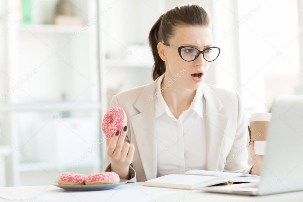 Indignant office manager with coffee and donut looking at display of laptop in front of her