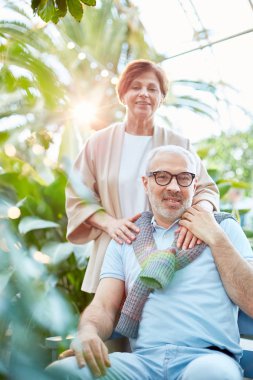 Mavi gündelik giysiler ve eşi arasında yeşil bitkiler doğal ortamda kamera bakarak mutlu olgun adam