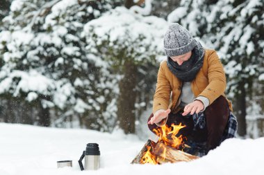 Genç adam kış gününde ormandaki rüzgârla oluşan kar yığını içinde otururken ateşte ellerini tutmak