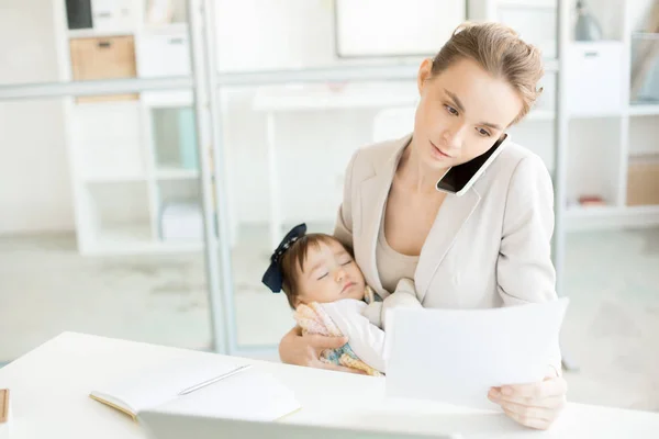 忙しい女性社員のスマート フォンで話して 彼女の腕の中で眠っている赤ん坊の娘ながらオフィスの重要なペーパーを通して見る — ストック写真