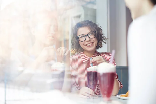 微笑的女孩在眼镜和条纹衬衣看她的朋友在谈话期间在咖啡馆里喝一杯 — 图库照片