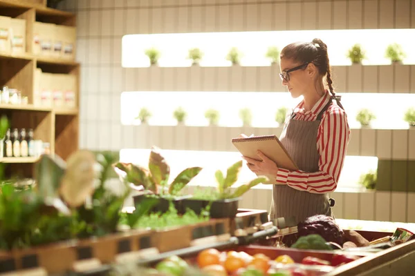 メモ帳でスーパーで販売のための植物の改訂をしながらノートを作る若い店員 — ストック写真