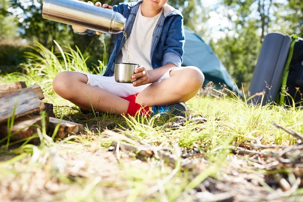 年轻的脱扣浇热水从热水瓶到他的杯子 而坐在草地上的篝火 — 图库照片