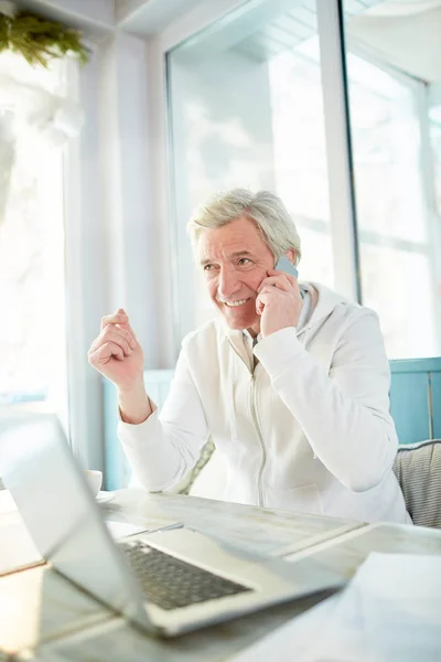 Modern senior man with smartphone calling his partner or colleague while sitting in cafe in front of laptop