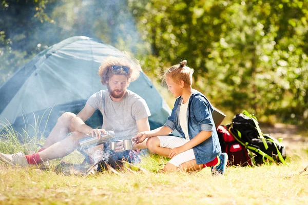 夏の日の喫煙キャンプファイヤーそば草の上に座って熱いお茶を持つ現代の若いスカウト — ストック写真