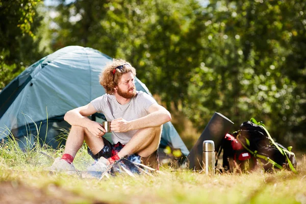 お茶のカップを押しながら芝生の上の背景に彼のテントに座っている普段着のまま穏やかな若いバックパッカー — ストック写真