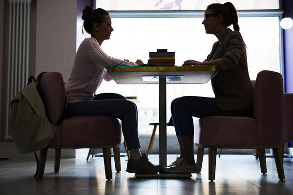 Silhouettes of two girls sitting opposite one another by table in cafe and having conversation