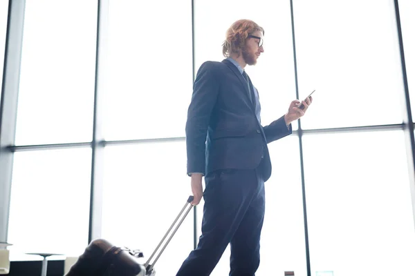 スマート フォンでメッセージングおよび飛行駅まで歩きながらスーツケースを引くエレガントなスーツの若い男 — ストック写真