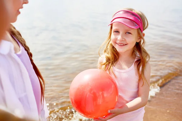 友人と余暇を過ごしながら水辺でカメラを見て赤い風船と幸せな愛らしい女の子 — ストック写真