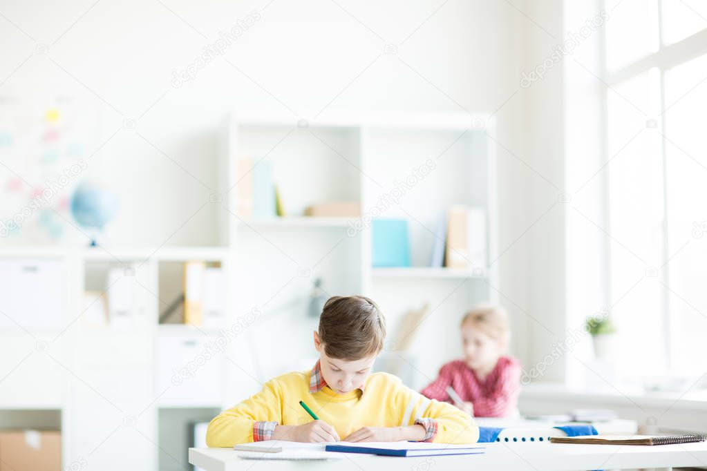 Serious schoolboy concentrating on written individual work by his desk at secondary school