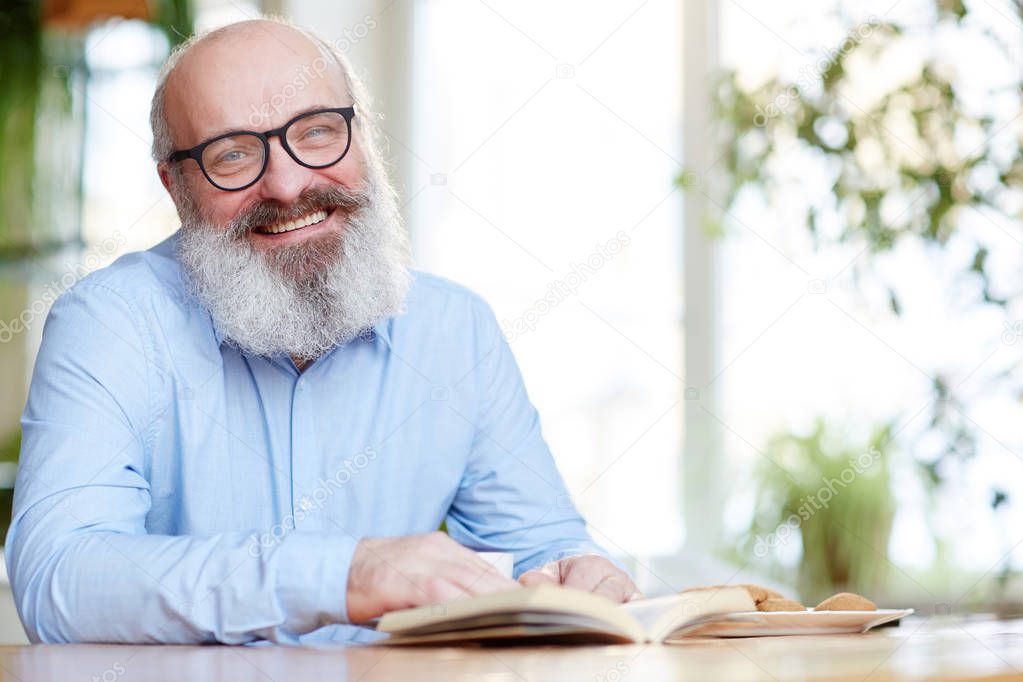 Smiling senior man in eyeglasses looking at camera while sitting by table with open book